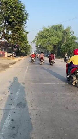 This video shows a line of motorcyclists on a paved road. The perspective is from one of the motorcyclists, looking forward and slightly to the right. The road is lined with trees on both sides, and there are buildings and a truck visible in the distance. The sky is clear and bright, suggesting it is daytime. The motorcyclists are all wearing helmets, and most are wearing jackets. They appear to be traveling in the same direction. The rider in the foreground is wearing a red helmet and a yellow jacket. The road has white lane markings, and there is a shadow of the rider and their motorcycle on the left side of the road.