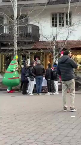 The video shows a street scene in a town with buildings and bare trees, possibly in a colder climate due to the clothing of people present. There is a group of people congregated around a table in front of a store. A person in an inflatable Christmas tree costume and another in an inflatable reindeer costume are also present. People walk by the gathering, some carrying shopping bags. The street is paved with light-colored stones. The general atmosphere suggests a festive, potentially holiday-themed event or promotion.