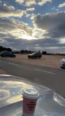 The video shows a partly cloudy sky with sunlight filtering through, illuminating a road and sandy area. A dark green Subaru Forester drives on the sand beside the road. Other cars are parked nearby. The camera is positioned inside a vehicle, with a pink coffee cup visible on the hood.