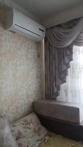 The media file shows an interior view of a room, likely a bedroom or living area, in Suklieia, Moldova. The scene is dominated by a large window covered with ornate, white lace curtains and heavy, draped pelmet curtains in muted tones of beige and grey. The curtains are elaborately styled, with a swagged top and a ruffled edge on the pelmet. The window itself is a broad, multi-paned structure, suggesting it could be a bay window.

To the left of the window, an air conditioning unit is mounted high on a wall covered with patterned wallpaper. The wallpaper has a subtle, textured damask design in cream and pale gold. Below the air conditioning unit, on a raised platform or window seat, sits a rectangular upholstered ottoman or cushion in a textured grey fabric. A pillow with a floral pattern rests on a surface in the foreground, partially obscuring a yellow textile, possibly bedding. The lighting suggests it is daytime, with natural light filtering through the lace curtains, creating a soft, diffused illumination. The overall impression is one of a well-decorated, albeit slightly dated, interior space. There is no visible text in the media file.