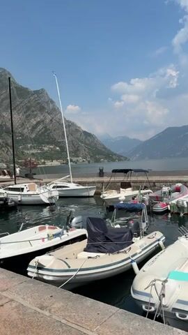 This video showcases a picturesque scene at a marina in Limone sul Garda, Italy. The primary subjects are various boats docked in the clear, turquoise waters of a lake, with a backdrop of imposing, mist-shrouded mountains. The time of day appears to be late morning or early afternoon, with a bright, sunny sky dotted with fluffy white clouds. The weather is pleasant and conducive to being outdoors by the water.

The video opens with a view of several inflatable dinghies and a sailboat, some with their covers on. As the camera pans, we see a long pier lined with more boats, including a larger white motor yacht named "LOVE IN BLUE" with Italian flags flying from its stern. The marina is bustling with activity, though no specific events are occurring. The general atmosphere is one of tranquility and leisure, with the natural beauty of the lake and mountains dominating the scenery.

Notable details include the detailed reflections of the boats and the sky on the water's surface. The architectural style of the surrounding buildings, visible in the distance, suggests a Mediterranean setting. Text is visible on one of the boats: "LOVE IN BLUE" on the stern of the motor yacht. The overall impression is one of a serene Italian lakeside town, perfect for a relaxing vacation or a day out on the water.