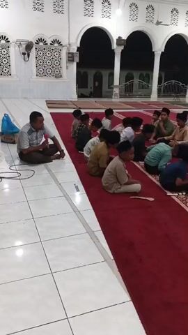 In a brightly lit, large indoor space with tiled flooring and red carpets, a group of young boys, dressed in various casual and traditional clothing, are seated in rows on the carpet. An adult male, wearing a light-colored collared shirt and dark pants, sits cross-legged on the tiled floor to the left of the boys, facing them. He is looking towards the boys and appears to be interacting with them. The boys are also seated cross-legged, facing the adult. Some boys are looking at the adult, while others are looking down or at each other. The architecture of the room suggests it may be a mosque or similar religious building, with arches and decorative elements visible. In the background, other people, including adults and possibly more children, are visible, some seated. The overall scene depicts an educational or religious gathering for the boys.