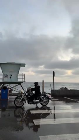 A person on a motorcycle drives past a lifeguard tower and the ocean on a cloudy day. The ground is wet with reflections of the scene.