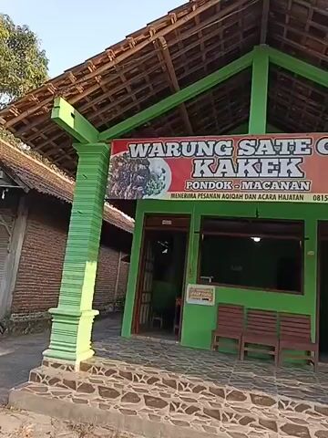 The video shows the exterior of a small, green building with a wooden roof. A banner above the entrance reads "WARUNG SATE GULE KAKEK". There are two red wooden benches outside the building. The building appears to be a restaurant. The ground in front of the building is paved with stones.
