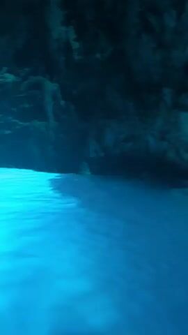 The video shows a view from a boat in a cave. The water is a bright blue, and sunlight is filtering in from an opening above, illuminating the water's surface. The cave walls are dark and rocky. In the foreground, a person is visible, holding up a phone to take a picture. The person appears to be sitting in the boat, and the camera is focused on the water and the cave.