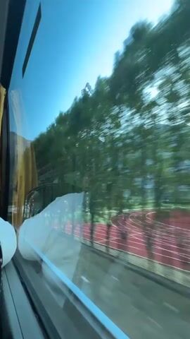 The video captures a view from inside a moving vehicle, looking out of a window. The initial frame shows trees and a running track outside, suggesting an urban or park-like environment. As the video progresses, the scenery shifts to reveal a mountainous landscape under a clear blue sky. A green car is visible in a parking lot, and a small white building is present. The mountains are large and imposing, with visible rock formations. The motion of the vehicle causes the trees and distant landscape to blur, creating a sense of speed and travel. The reflection of the interior of the vehicle, including a yellow curtain and what appears to be a white blanket or pillow, is also visible on the window. A street lamp is seen passing by in the later frames.