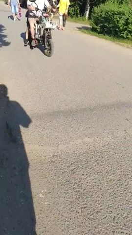 The video opens with a man in a bucket hat riding an electric scooter with a child standing on the back. They are moving away from the camera on a paved road. In the background, another person walks, and two individuals ride horses. The scene then transitions to a child wearing a white helmet and a dark t-shirt riding a light brown horse, led by a woman in a yellow t-shirt walking beside it. They are moving towards the camera on the same paved road, with trees and a fence visible in the background.