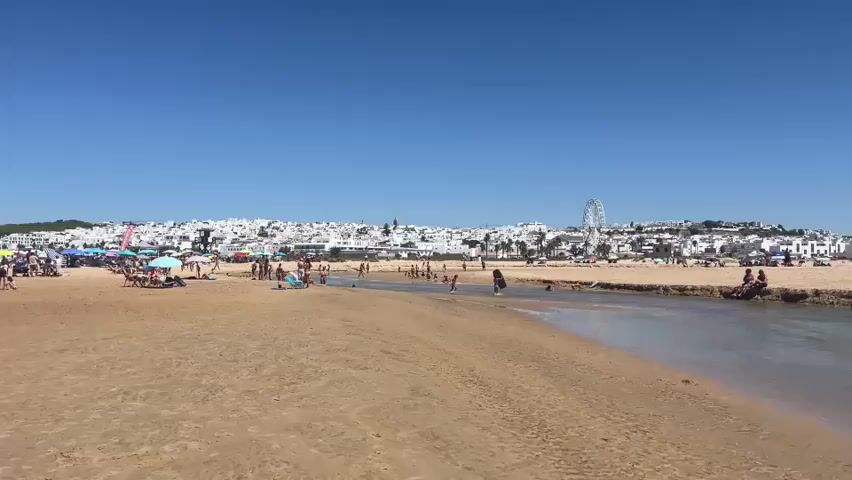 A sunny day at a beach with a river running through it. The beach is sandy with some people sitting on chairs under umbrellas and others walking along the shore. There are people in the river, some swimming and others wading. In the background, there is a white-washed town with buildings on a hill and a Ferris wheel. The sky is clear and blue.