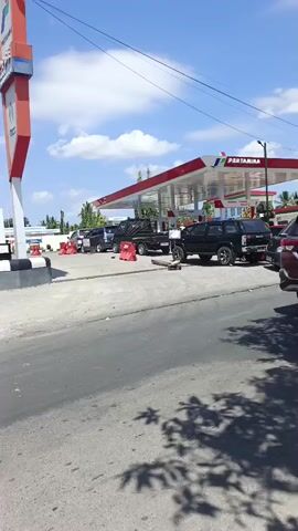 The video shows a view of a Pertamina gas station and the surrounding street. Several vehicles, including trucks, SUVs, and a motorcycle, are visible at the gas station and on the road. Some vehicles appear to be queuing. In the background, there are buildings, palm trees, and a blue sky with clouds. An Alfamart store sign is also visible. The overall impression is of a typical street scene in a sunny environment.