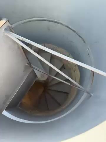 The video begins with a view looking down a narrow, light-colored spiral staircase with modern metal railings. The camera then moves upwards, revealing a panoramic cityscape bathed in bright sunlight under a clear blue sky. Numerous buildings with tiled roofs and varying architectural styles stretch across the urban landscape, with distant hills or mountains on the horizon. The view then pans further to reveal the vast expanse of the Mediterranean Sea, dotted with several large ships. The foreground shows a paved outdoor viewing area with a low stone wall, overlooking the city and the sea. The sun is high in the sky, indicating a bright day. No people are visible in the video.