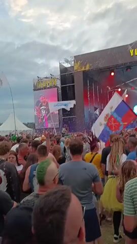 The video shows a large crowd of people at an outdoor concert. Many people are holding up their hands, some with fists raised in a rock-and-roll gesture. A Russian flag is prominently displayed in the foreground. The stage has a large screen showing graphics and lights. There are also several flags visible in the background. The sky is cloudy. The atmosphere appears to be energetic and celebratory.