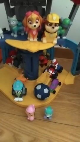 The video shows a toy play set that appears to be a lookout tower. There are several toy figurines of dogs dressed in various uniforms and outfits, which are likely characters from the Paw Patrol franchise. Some of the dogs are positioned on different levels of the tower, while others are scattered around the base. There are also other toy vehicles and accessories visible, such as a blue and white soccer ball and several small toy cars. The play set is situated on a wooden surface, possibly a floor or table, with a wall in the background. The lighting is dim, and the camera is slightly out of focus, giving the video a somewhat hazy appearance.
