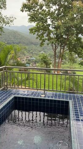 The video shows a small tiled plunge pool with rippling water on a balcony or patio. The pool is surrounded by a metal railing. Beyond the railing, there is a lush green landscape with rolling hills and trees. In the distance, a small village or cluster of houses can be seen nestled among the trees. The sky is overcast.