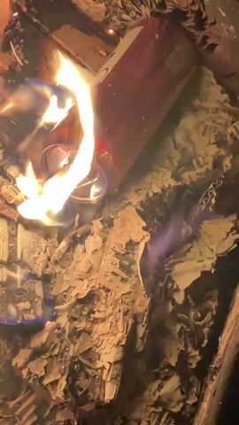 The video captures a close-up of a red and white aluminum beverage can engulfed in flames within a burning pit. Bright yellow and blue flames are visible, consuming what appears to be paper or wood ash surrounding the can. The can itself is deforming from the intense heat, with the top portion and opening mechanism glowing. The background is dark, suggesting the scene takes place at night or in low light. Russian speech can be heard in the background. The location is Веревское, Russia.