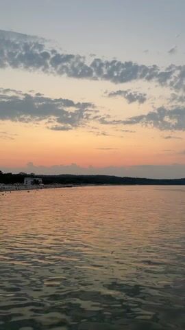 A wide shot captures a serene lake at dusk. The sky is a gradient of pale blue to soft orange, with scattered clouds. The water reflects the sky's colors, appearing calm and rippling gently. In the distance, a shoreline is visible, lined with trees and a few buildings. There are small figures of people on the shoreline, appearing as distant specks against the landscape. The overall impression is one of peaceful transition from day to night.