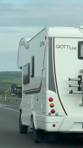 The video shows the side of a white Giottiline motorhome driving on a highway. We see the windows, taillights, and badging of the vehicle as the camera passes it. The background shows a green, hilly landscape under a cloudy sky. The camera briefly focuses on the view out the window of the car filming.
