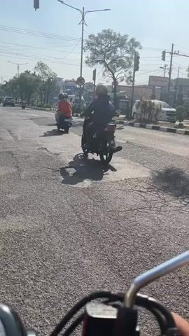 The video shows a view from a motorcycle on a sunny day, looking ahead at a road with several other motorcycles. The road surface is asphalt and appears to be in poor condition with visible cracks and patches. Two motorcycles are ahead, one with a rider wearing a white shirt and dark pants, and another behind it. Traffic lights and street signs are visible on the right side of the road. Trees and buildings are present in the background. The perspective suggests the viewer is also on a motorcycle, with a portion of the handlebars visible at the bottom of the frame.