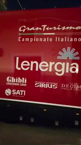 The video shows a red truck with white text on its side. The text includes "Gran Turismo" and "Campionato Italiano". Below that, larger text reads "Lenergia" with a logo above it. Several smaller logos are also visible, including "Chibli", "SATi", "SIRIO", and "DECIM". The truck is on a street at night, illuminated by streetlights and lights from nearby buildings. The street appears to be paved with cobblestones. People are visible in the background, walking along the street. The truck is moving slowly.