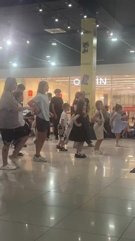 A group of children and adults are performing a dance routine in a public indoor space, possibly a shopping mall. The floor is polished and reflects the overhead lights, creating a glossy appearance. In the background, large glass windows reveal shops with displays. A tall, yellow pillar with images of cats and text is visible. The individuals are positioned in a line, facing the camera, and appear to be following choreographed movements. One young girl in a black polka-dot dress is in the foreground, with her hands on her hips. Other children are in various positions, some with one leg raised. The lighting is bright, casting shadows on the floor.