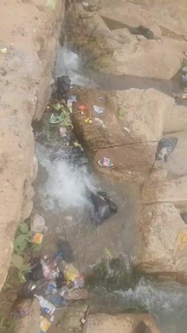 A drone flies over a polluted stream. The water is murky and filled with trash. Plastic bags, bottles, and other debris are scattered along the banks and in the water. The stream flows through a rocky, overgrown area. The overall impression is one of neglect and environmental damage.