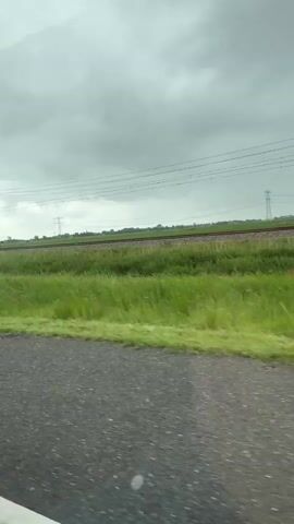 The video shows a view from a vehicle moving along a paved road. To the left, the side mirror of the vehicle is visible. Beyond the road is a field of tall green grass, with a railway track running through it. Overhead, there are power lines and poles supporting them. The sky is overcast with grey clouds. The scene appears to be a rural or semi-rural landscape.