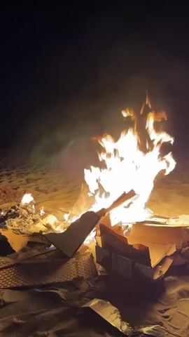 This media file depicts a nighttime scene on a sandy beach, likely Playa Del Sol-Villacana, Spain. The central subject is a bonfire, with large, bright flames consuming cardboard boxes and other debris. The fire is intensely bright, casting a warm orange and yellow glow on the surrounding sand and smoke. The sand is visible in the foreground and background, textured by what appears to be footprints or the natural contours of the beach.

The time of day is clearly nighttime, indicated by the dark sky. There are no people visible in the frame. The activity taking place is the burning of materials in a campfire setting. The cardboard boxes are stacked and some pieces are already partially burned, with edges blackened and warped.

Notable details include the smoke rising from the fire, creating a hazy effect against the dark sky. The overall mood is one of warmth and perhaps a sense of catharsis or simple enjoyment of a fire on the beach. The lighting is dramatic due to the intense flames, with deep shadows in the less illuminated areas. There is no visible text within the media file.