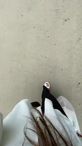 The video is a top-down shot of a person walking on a concrete surface. The person is wearing white sandals and black pants. They are also carrying a white plastic bag in their right hand. The bag appears to contain some items. The concrete surface has some cracks and stains. There is some greenery visible on the right side of the frame. The person's hair is visible in the foreground.