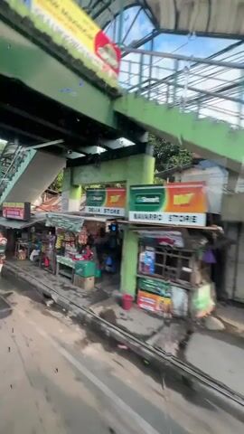 The video shows a street scene with a pedestrian overpass and several small shops lining the street. A truck is parked nearby, and people are seen interacting with the shops and the truck. The environment appears to be urban, with trees and power lines visible. The weather seems to be fair with a blue sky and some clouds.