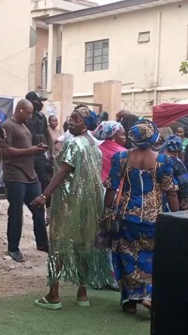 The video captures an outdoor social gathering in Sabon Gari West, Nigeria, under bright daylight. A diverse group of adults is seen mingling and interacting.

In the foreground, an artificial green turf covers the ground, transitioning to dirt and debris further back. A prominent figure is a woman wearing a dazzling, sequined light green kaftan with fringed details, a blue headwrap, and light green sandals. She smiles broadly and gestures with her hands, appearing to dance or express joy in response to the music playing in the background.

To her left, a man in a green short-sleeved collared shirt and dark trousers, possibly event staff, observes the scene. Another man in a brown t-shirt, dark trousers, and glasses is seen looking at his phone while holding what appears to be a microphone. A woman in a blue and gold patterned traditional dress and headwrap is also present, looking towards the woman in green. Other attendees, including a woman in a pink top, are visible in the background.

The setting includes a light-colored building in the background. A banner featuring a person's portrait is partially visible on the far left. A white sign with the number "21" is also visible in the background. The overall atmosphere appears festive and lively, with people enjoying themselves.