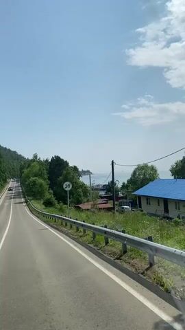 The video shows a car driving on a paved road. The road curves to the left, and there is a guardrail on the right side of the road. There are trees and vegetation on both sides of the road. In the distance, there is a body of water and some buildings. The sky is blue with some clouds. There is a speed limit sign with "20" on it. Several other cars are visible on the road. The video appears to be shot from inside a moving vehicle.