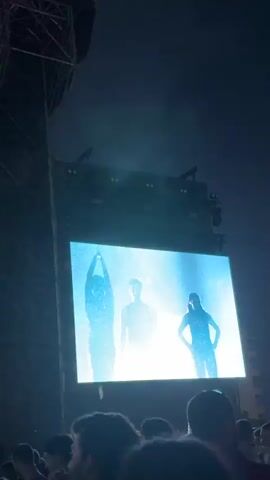 A video captures a concert stage at night, starting with a screen displaying a vaguely humanoid figure adorned with illuminated shapes. The camera then pans upward, revealing the scaffolding, lighting equipment, and fog machines above the stage. The name "LA GOSSA SORDA" is prominently displayed at the top of the structure. The shot ends with the stage in near darkness. The heads of audience members are visible in the foreground.
