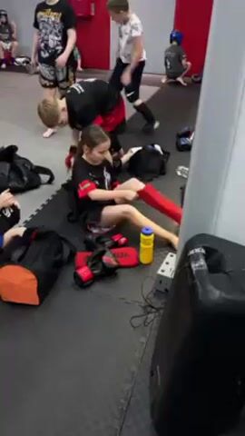 Several young people are sitting on a black floor mat in a room with red and grey walls. Most are putting on or taking off boxing equipment. A young girl wearing red shin guards is sitting on the mat, along with red boxing gloves, a yellow water bottle, and several black duffel bags. A boy is helping her with something in one of the bags. Other children are putting on boxing gloves and getting ready for a class. One person is sitting against the wall wearing a blue boxing helmet. A large speaker is in the foreground.