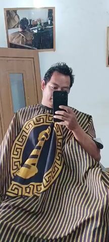 A man is sitting in a barber chair, wearing a haircut cape. He is holding a smartphone in his right hand, taking a selfie. The cape is striped brown and yellow, with a large oval design in the center featuring a golden horn-like object within a Greek key pattern. The man has dark, slightly messy hair and is looking at his phone. In the background, a mirror reflects the scene, showing the man from behind. There is also a wooden door and a light-colored wall. The overall setting appears to be a barber shop.