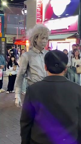 The video shows a street performance in what appears to be a city in South Korea. A person dressed in silver paint and costume with long, silver hair is the performer. He is making sudden movements and interacting with a man in a black jacket and black cap who is facing him. Behind them, a crowd of young women and other pedestrians observe and react with amusement. The scene is set on a pedestrian street with shops and signs in Korean visible in the background. The lighting suggests it is either late afternoon or early evening.