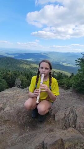 A young girl with braided brown hair, wearing a bright yellow t-shirt and dark shorts, kneels on a rocky outcrop. She is playing a white recorder. Her expression is focused as she looks directly at the camera. Behind her, a vast landscape unfolds, characterized by rolling green hills and mountains stretching towards the horizon under a vibrant blue sky dotted with fluffy white clouds. The scene is bathed in natural daylight, suggesting it is daytime. The lush vegetation and the distant mountain ranges hint at a natural, possibly mountainous, setting. The presence of pine trees on the right side of the frame further reinforces this impression of a wilderness or park-like environment. The overall mood is peaceful and serene, with the girl's musical activity adding a touch of quiet joy to the majestic scenery. The location is identified as Sutton, Canada.