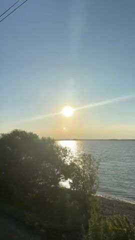 The video opens with a shot of a lake under a bright, sunny sky. Trees are visible on the left side of the frame, and a shoreline stretches along the right. As the video progresses, a white car is seen parked on the beach with its trunk open. A person is standing next to the car, appearing to unpack or prepare fishing gear. There is a fishing rod set up nearby. The scene depicts a calm, outdoor setting, possibly a recreational trip to the lake.
