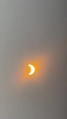 The video shows a solar eclipse. The sun is partially obscured by the moon, creating a crescent shape. The sun is a bright orange color, and the sky is a hazy gray. The footage is grainy and appears to be captured on a mobile phone. The video is brief, lasting only a few seconds.
