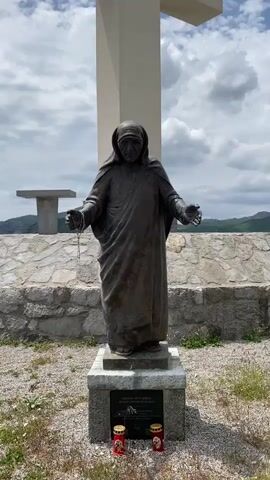 The video features a slow zoom-in on a bronze-toned statue of Mother Teresa, standing outdoors. She is depicted in her characteristic habit, with both hands extended forward, holding rosary beads draped between them. Behind the statue is a large, prominent white cross.

The statue rests on a multi-tiered stone base. A plaque is affixed to the front of the base, with visible text including "SHENJTA NËNË TEREZA" (Saint Mother Teresa) and "ANJEZË GONXHE BOJAXHIU" (Agnes Gonxha Bojaxhiu), her birth name, with additional smaller text or dates below. Small red votive candles are placed at the very bottom of the base.

The setting appears to be an elevated outdoor location, possibly a hilltop, offering views of distant mountains or hills. A low stone wall or barrier is directly behind the statue, and a simple, flat concrete table or bench is visible further in the background to the left. The sky is partly cloudy with patches of blue, suggesting bright daytime weather. The scene conveys a solemn and reverent atmosphere at this religious monument in Benkalj, Montenegro.