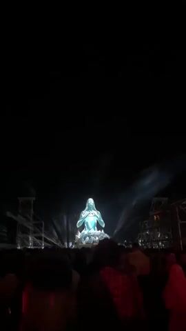 A large, illuminated, statue-like figure of a person in a meditating pose sits on a platform in the center of an outdoor nighttime event. The figure is primarily blue and white, with bright lights emanating from it and projecting outwards in beams. A crowd of people is visible in the foreground, silhouetted against the stage lighting. The environment appears to be a festival or concert, with stage lighting structures visible on the sides. The camera is positioned among the crowd, looking towards the stage. The video pans slightly upward and includes bursts of light and some atmospheric haze or smoke.