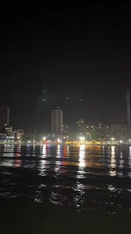 A nighttime view of a city skyline reflected in the water. The city is illuminated by numerous lights, including those from tall buildings and a waterfront area that appears to be decorated with string lights. The water is choppy, with the lights creating shimmering reflections. The sky is dark and there are some faint green lights visible in the distance, possibly from a bridge or other structure.