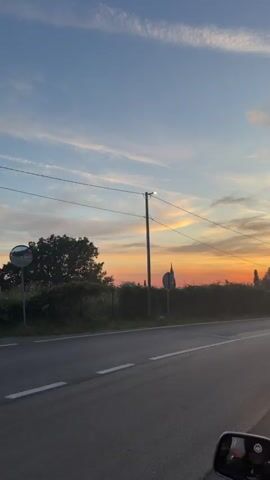 The video shows a sunset view from a moving vehicle. The sky is painted with hues of orange, yellow, and blue, with wispy clouds. A paved road with a white line runs through the frame, bordered by a dense green hedge and trees. A utility pole with wires stands on the left, and a road sign is visible behind the hedge. In the distance, a tall, thin structure, possibly a tree or a tower, pierces the horizon. As the vehicle moves, a black car parked on the side of the road comes into view, followed by more trees and a glimpse of what might be a rural landscape. A rearview mirror is visible at the bottom right corner of the frame.