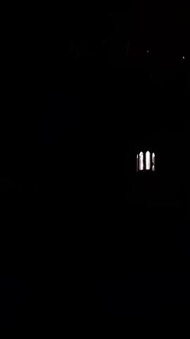 The video captures a nighttime scene of fireworks exploding in the sky above a dark silhouette of a building with illuminated arched windows. The first firework is red, followed by a circular green firework. The building appears to be a church or a similar structure. The video is shot from a low angle, looking upwards at the sky.
