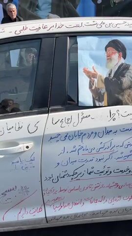 A white car is parked on an asphalt surface, with its doors covered in handwritten text and images. On the driver's side door, a photograph of a man with a beard and a turban is affixed, his hands clasped in prayer. Below this image, more handwritten text is visible, including some phrases in a larger script. A dark-colored car door handle is present, and there are reflections on the window. The car is positioned in front of a large banner displaying images of men, possibly political figures. Other people are partially visible in the background, including one individual looking towards the camera. The overall impression is of a display or demonstration.
