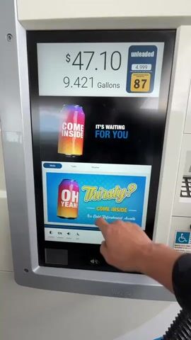 The video shows a close-up of a gas pump touchscreen display. The top of the screen shows the price in dollars and the number of gallons dispensed. The middle section of the screen displays advertisements for beverages, with cans of soda and text that changes between "COME INSIDE FOR YOU" and "OH YEAH! Thirsty? COME INSIDE Ice Cold Refreshment Awaits". The bottom section of the screen displays a map with traffic information. A person's hand is seen interacting with the touchscreen, pressing various buttons and sections of the display. The background is blurred but appears to be an outdoor environment, possibly a gas station. The ARCO logo is visible at the bottom of the screen.