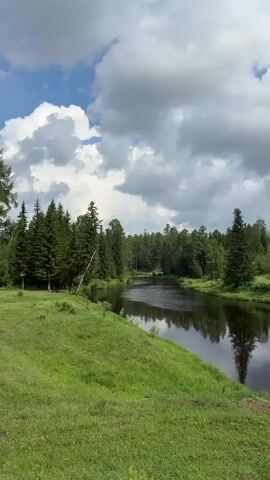The video shows a river winding through a lush green landscape under a cloudy sky. On the far bank of the river, trees and a small village with houses and a bridge can be seen. The foreground features a grassy slope and a rocky area. The camera pans slowly to the right, revealing more of the village and the surrounding hills. There are no people visible in the video.