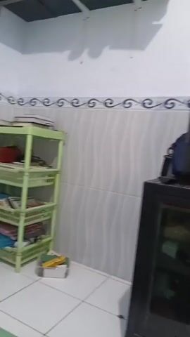 A panning video captures a corner of a room. On the left is a light green, four-tiered plastic shelf unit filled with books, folders, and other items. To the right of the shelf is a black cabinet with a glass door, topped by a television. The walls are white with a tiled border near the ceiling, and the floor is tiled in white squares.