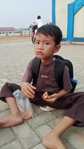 A young boy, appearing to be of elementary school age, sits on a paved outdoor area with a black backpack on. He is wearing a dark brown t-shirt and pants. His legs are spread wide in front of him, and he is barefoot. In his right hand, he holds a piece of food, and his left hand is near a clear plastic bag filled with red items. He looks directly at the camera with a pouting expression. In the background, there are several motorcycles, people, and a building with blue trim. Another child, wearing a red shirt, blue jeans, and a light-colored hat and hijab, is visible in the background, walking away from the camera and holding a piece of food. The overall environment appears to be an open, possibly school-like, outdoor space with paved walkways and some dry grass.