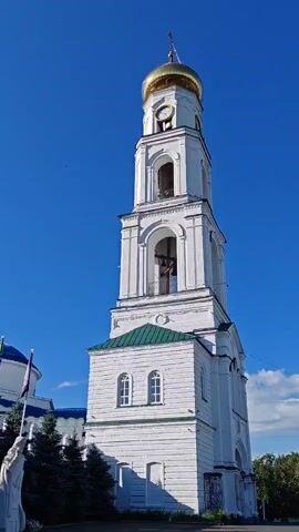 The video shows a tall, white bell tower with a golden dome against a clear blue sky. There are trees in the foreground, and a glimpse of another building with a blue dome on the left side. The bell tower has multiple levels with arched openings and windows. The clock face on the tower is visible. The sound is of a bell ringing.