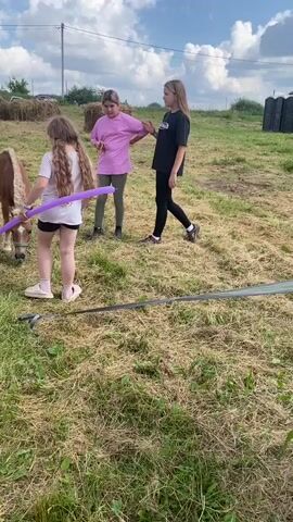 The video begins with a group of three young people, two girls and a boy, in an outdoor grassy area. The girl on the left, with long brown hair, is holding a long purple balloon and interacting with a small brown pony. A second girl, standing behind her, is wearing a pink shirt and dark pants. A third girl, on the right, wears a black t-shirt and black leggings. The boy, wearing a yellow t-shirt and a green cap, stands to the left of the pony and holds a yellow balloon. The background shows hay bales, trees, and a cloudy sky. The camera pans to the right, revealing more of the grassy field and a large inflatable structure with red and blue sections. Several people are visible near tents and hay bales in the distance. The video then cuts to a scene under a canopy where a woman dressed as a unicorn, holding a pink balloon, is interacting with four young girls. A person in a yellow mascot costume stands to the left. The ground is covered in grass.