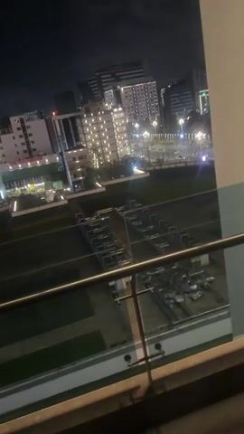 The video captures a night panoramic view of a city, likely Addis Ababa, Ethiopia, from a high vantage point, possibly a balcony. The camera pans slowly from left to right, revealing a vibrant urban landscape illuminated by artificial lights.

Numerous buildings of varying heights and architectural styles are visible, with many windows lit up, suggesting residential or commercial activity. Streetlights cast a glow on roads, and streaks of light from moving vehicles can be observed due to motion blur, indicating traffic. In the foreground, a modern balcony railing made of metal and glass is prominent. Below, there appear to be landscaped green areas or parks, also partially illuminated. The sky is dark, characteristic of nighttime, with no visible stars or clouds. No people or specific activities are distinctly visible, beyond the general hustle of a city at night. There is no discernible text within the video.